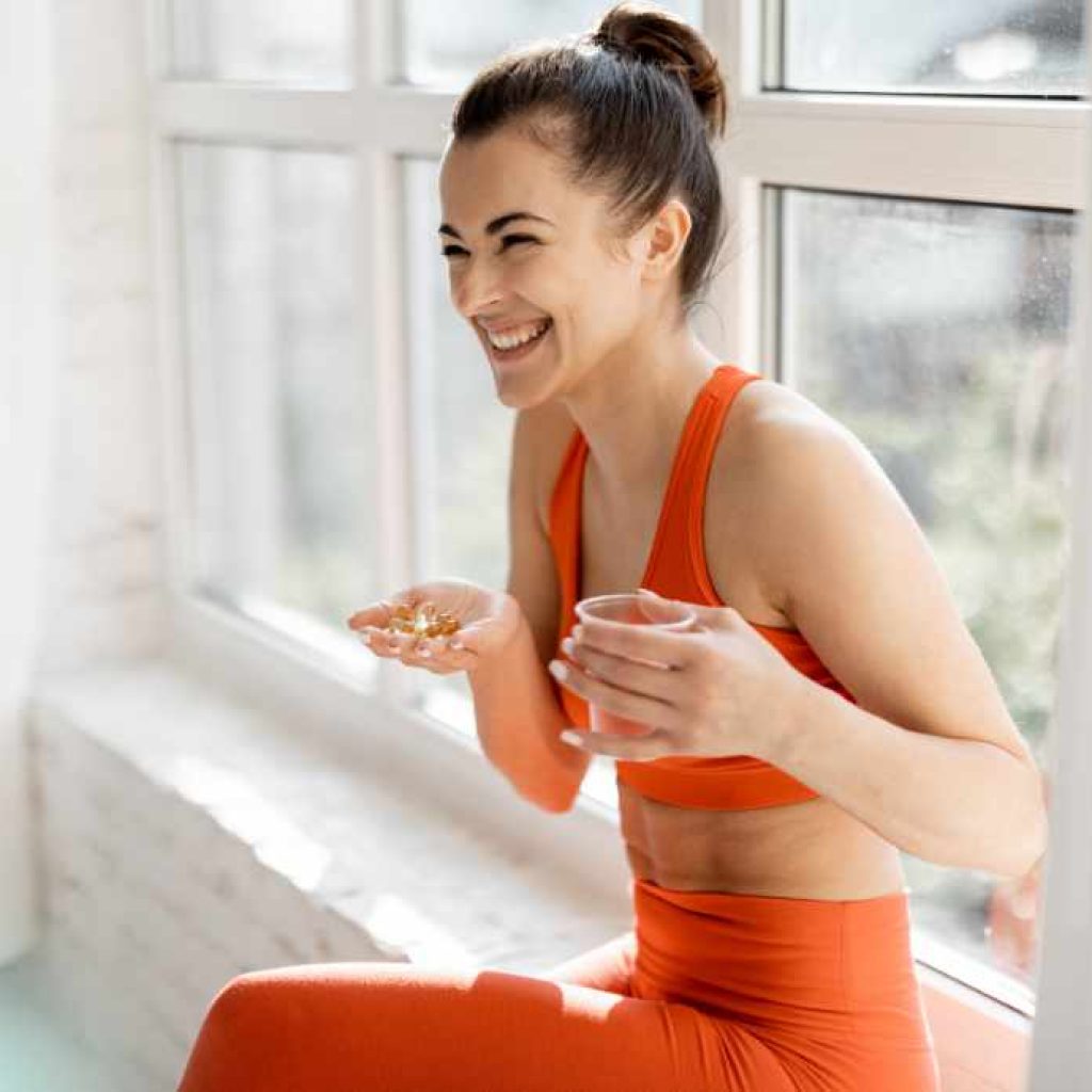 Woman on window sill with vitamins and water in her hands smiling