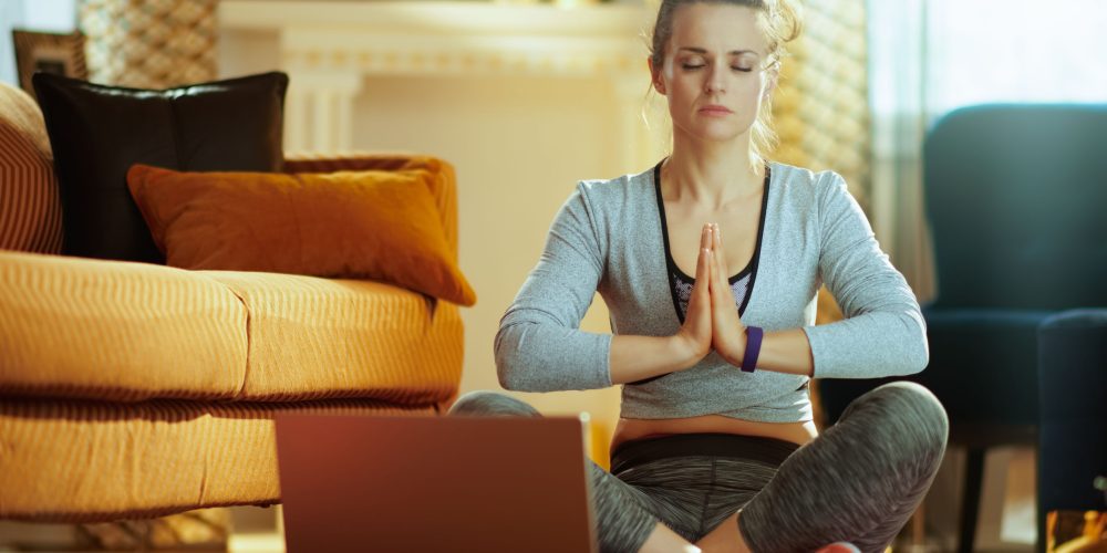 relaxed fit sports woman in fitness clothes in the modern house meditating using online yoga training program in laptop.